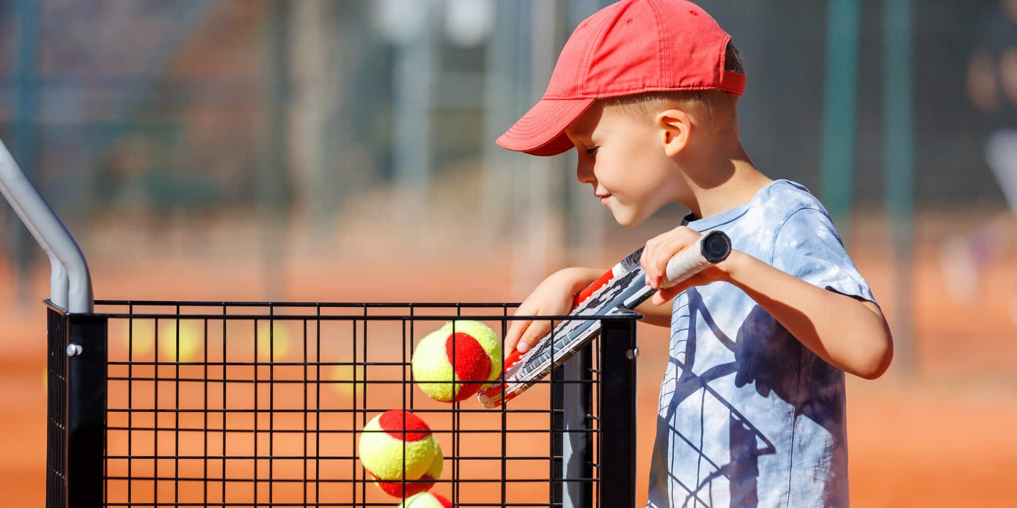 Cute little happy kid boy enjoy collecting balls in basket cart sign plate Kids-training clay tennis court at lesson. Summer sport camp. Sports youth school. Healthy child kid active lifestyle.